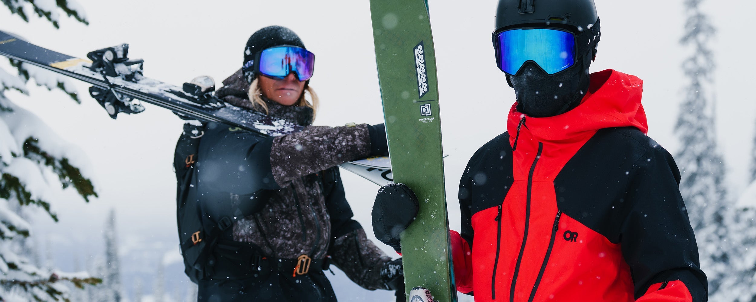 Two friends holding their skis in the foggy snow wearing Outdoor Research Men's Powderverse Jacket Ranger Green Granite Print/Black and Lingonberry/Black. 