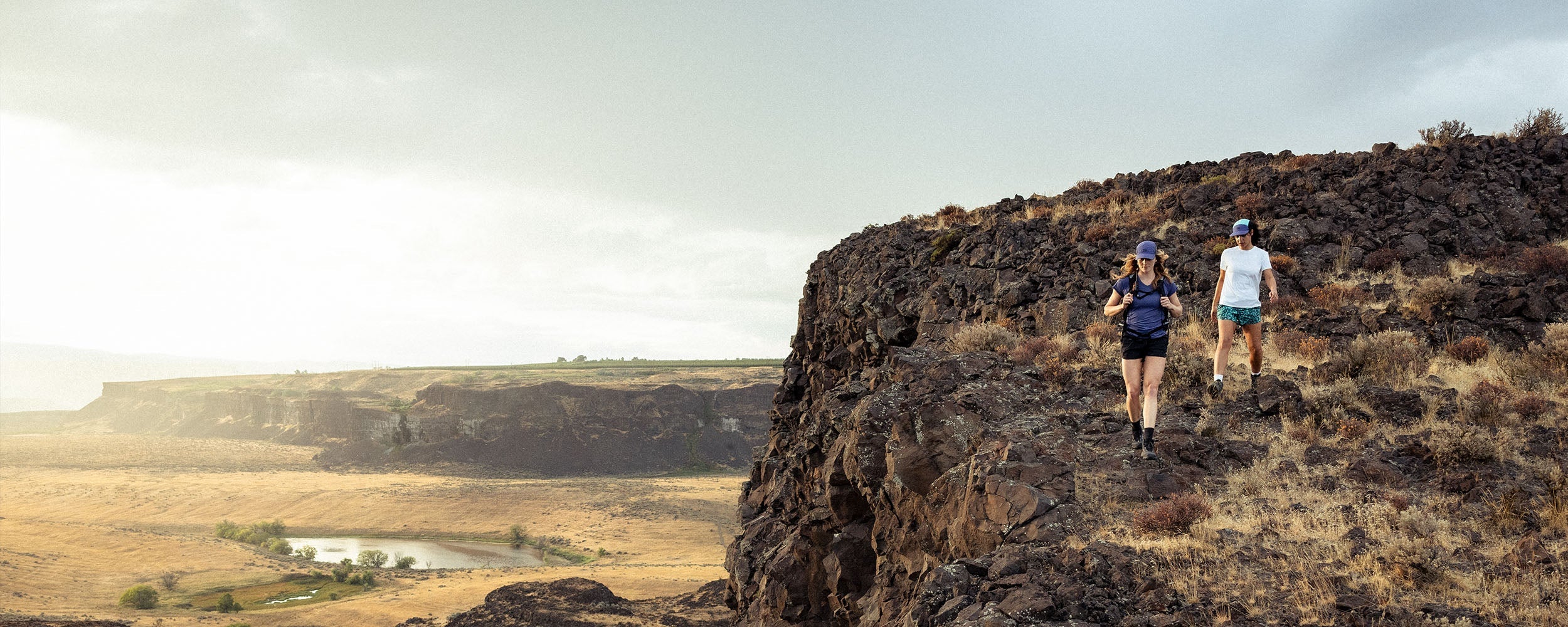 Two friends walk along the ledge of a rock cliff with a desert plateau behind them.