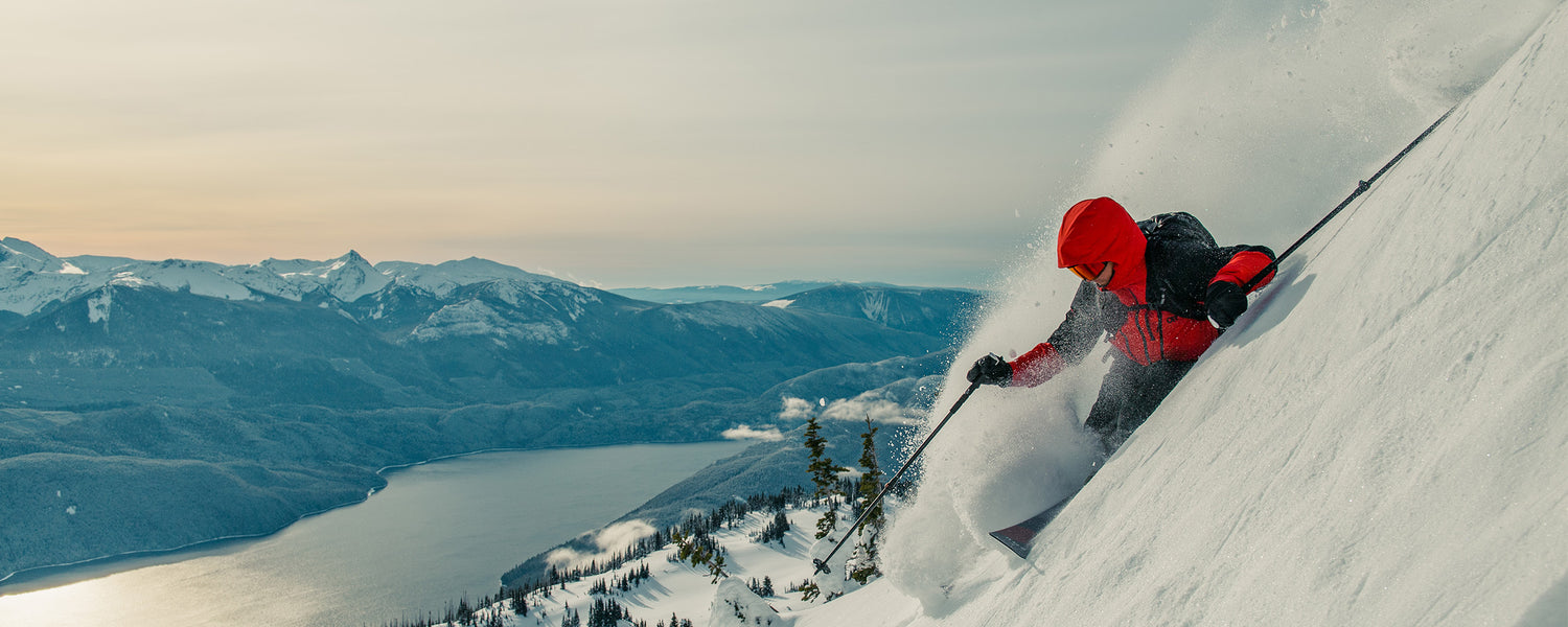 Skier going down steep mountain with a view of the water wearing Outdoor Research Men's Powderverse Jacket Lingonberry/Black. 