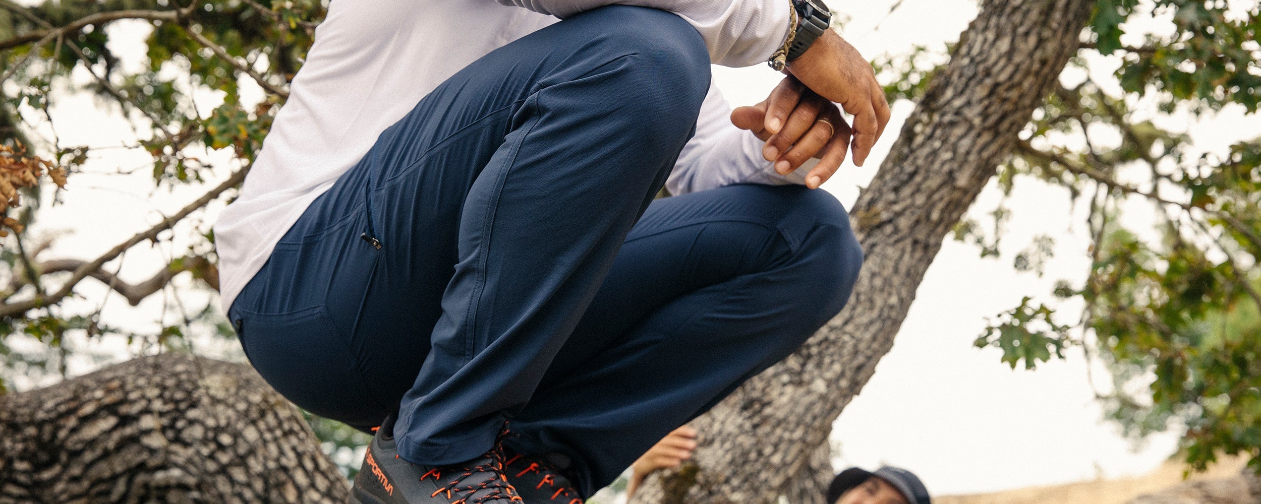 Man crouches on a branch of a tree he climbed while wearing Outdoor Research Ferrosi Pants in Dark Navy.